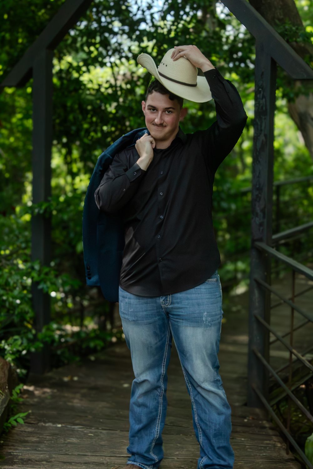 A person wearing a cowboy hat, black shirt and jeans poses on a wooden bridge surrounded by green foliage.