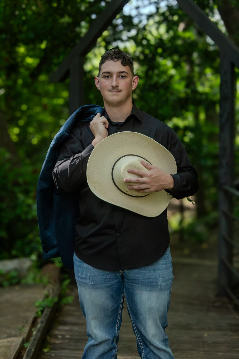 A person in a black shirt and jeans holds a cowboy hat while standing on a wooden bridge surrounded by green foliage.