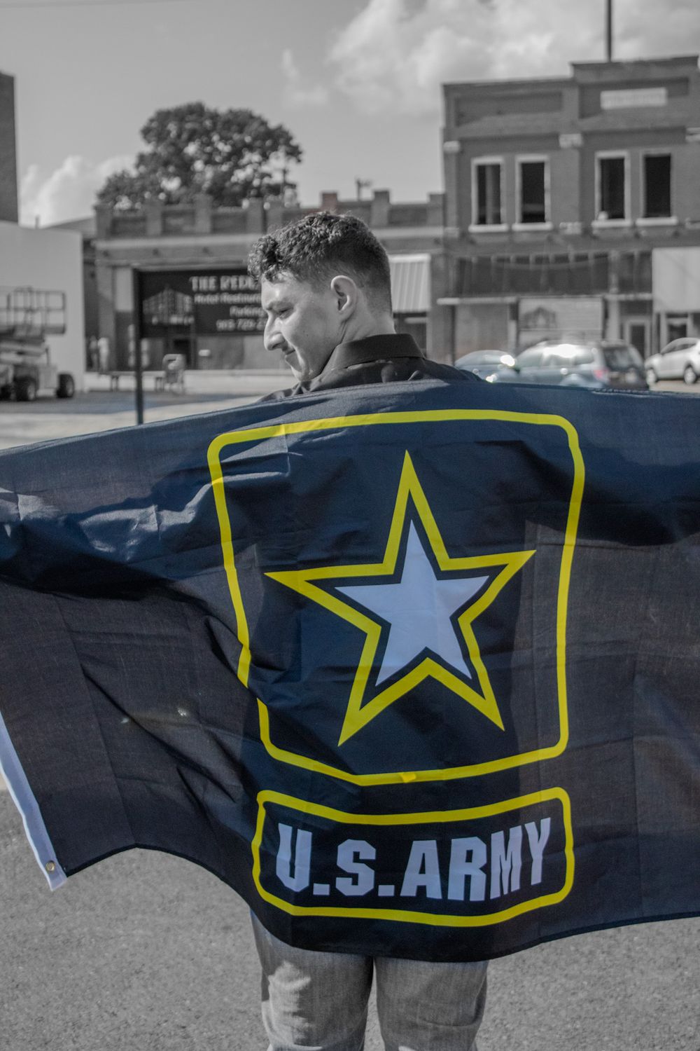 Person holding up a black flag with U.S. Army logo and star emblem in front of small town storefronts.