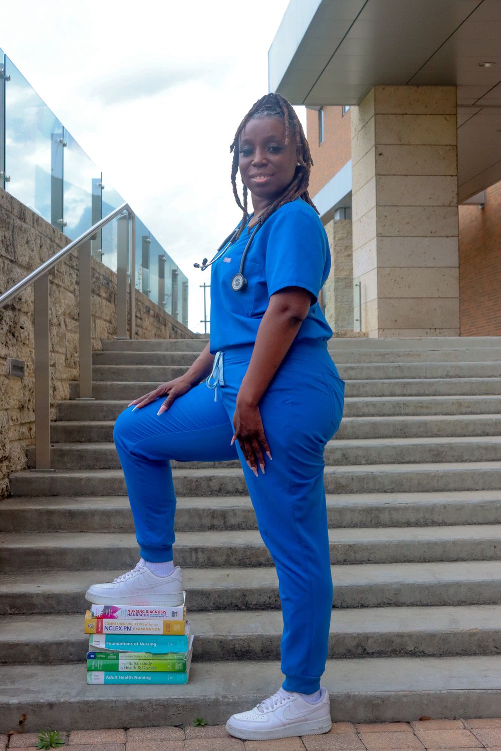 A nurse in blue scrubs with a stethoscope poses on steps with medical textbooks outdoors.