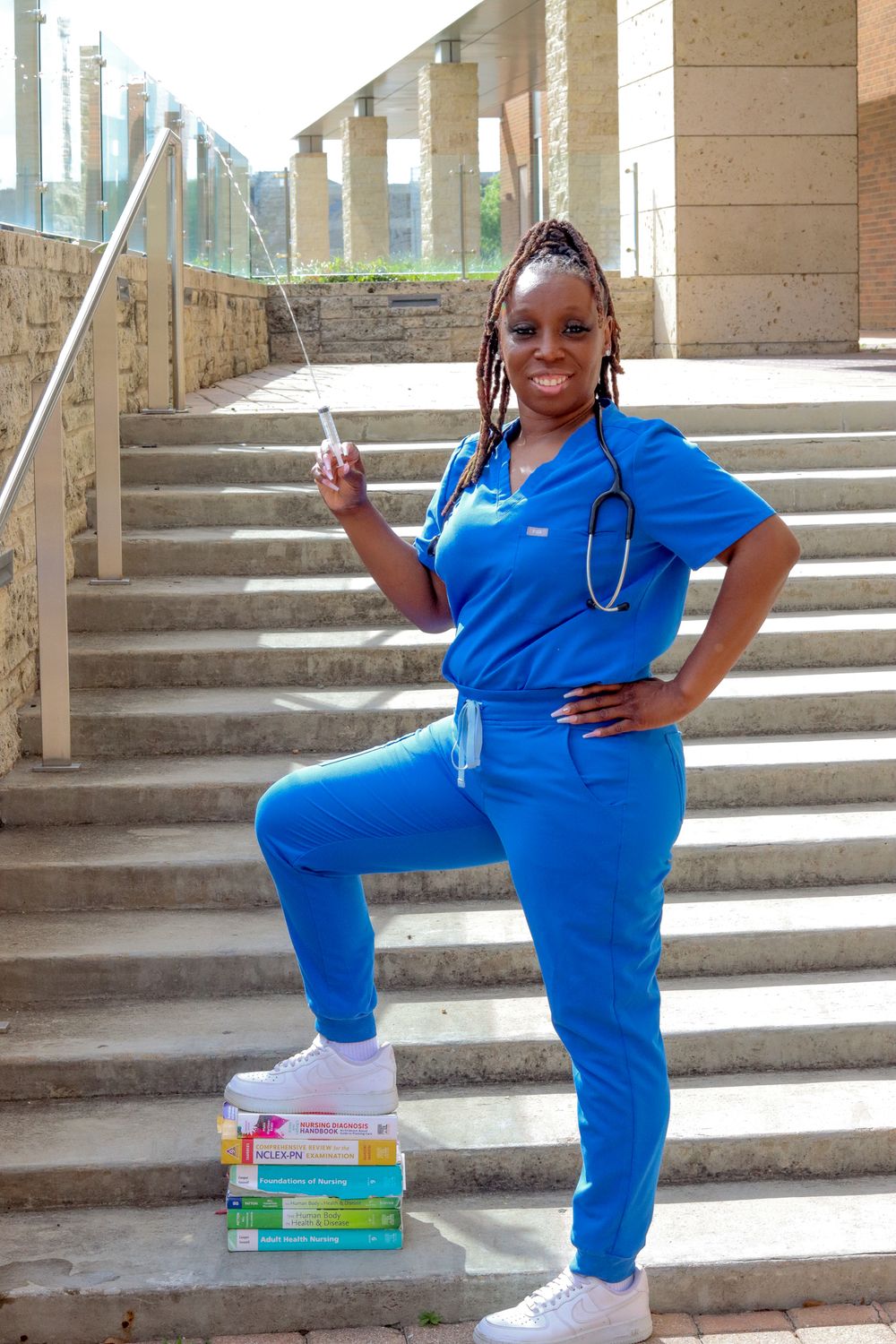 A Black female nurse in blue scrubs poses on stairs with medical books, stethoscope, and syringe.