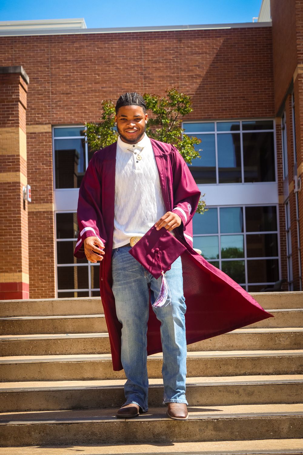 Young man in maroon graduation gown walks down school steps, smiling and holding cap on sunny day.