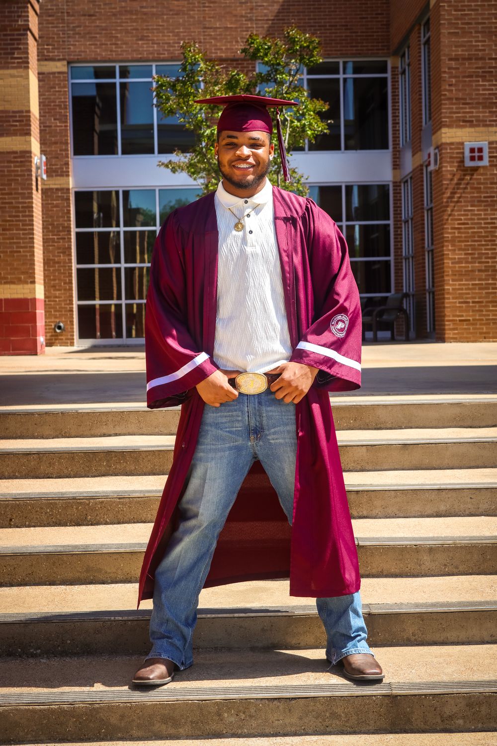 Young man in maroon graduation cap and gown smiling on steps outside a brick building.