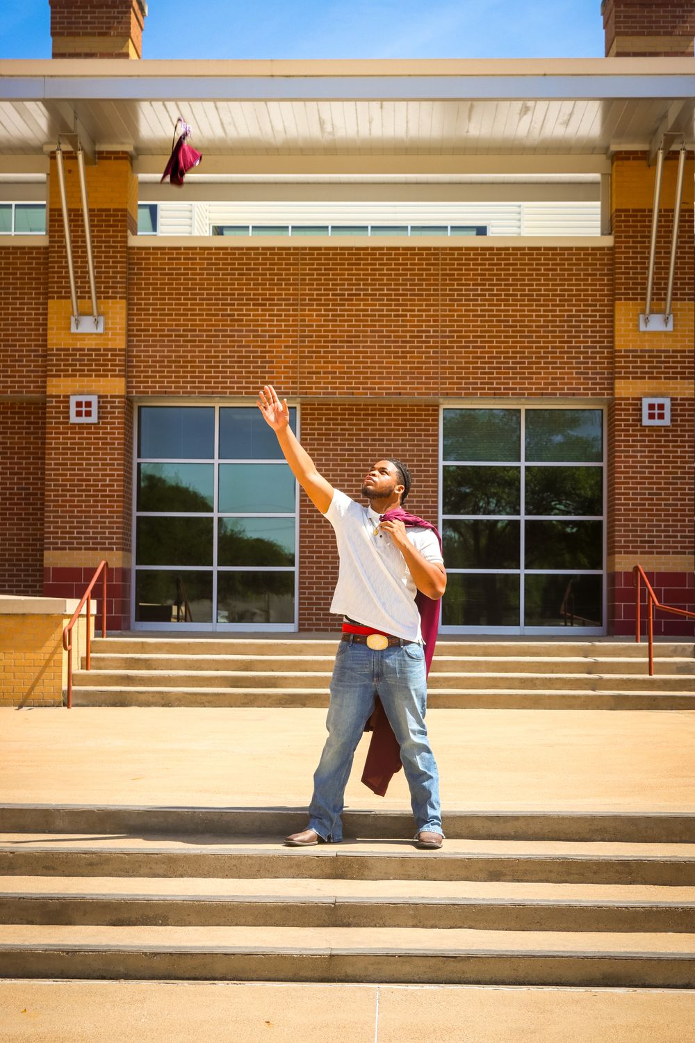 A graduate in jeans tosses his maroon cap in the air outside a brick university building on a sunny day.
