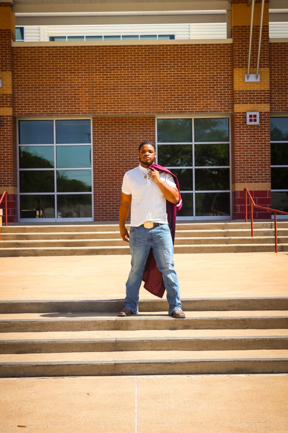 A young man in jeans and white shirt holds a maroon graduation gown over his shoulder on school steps.