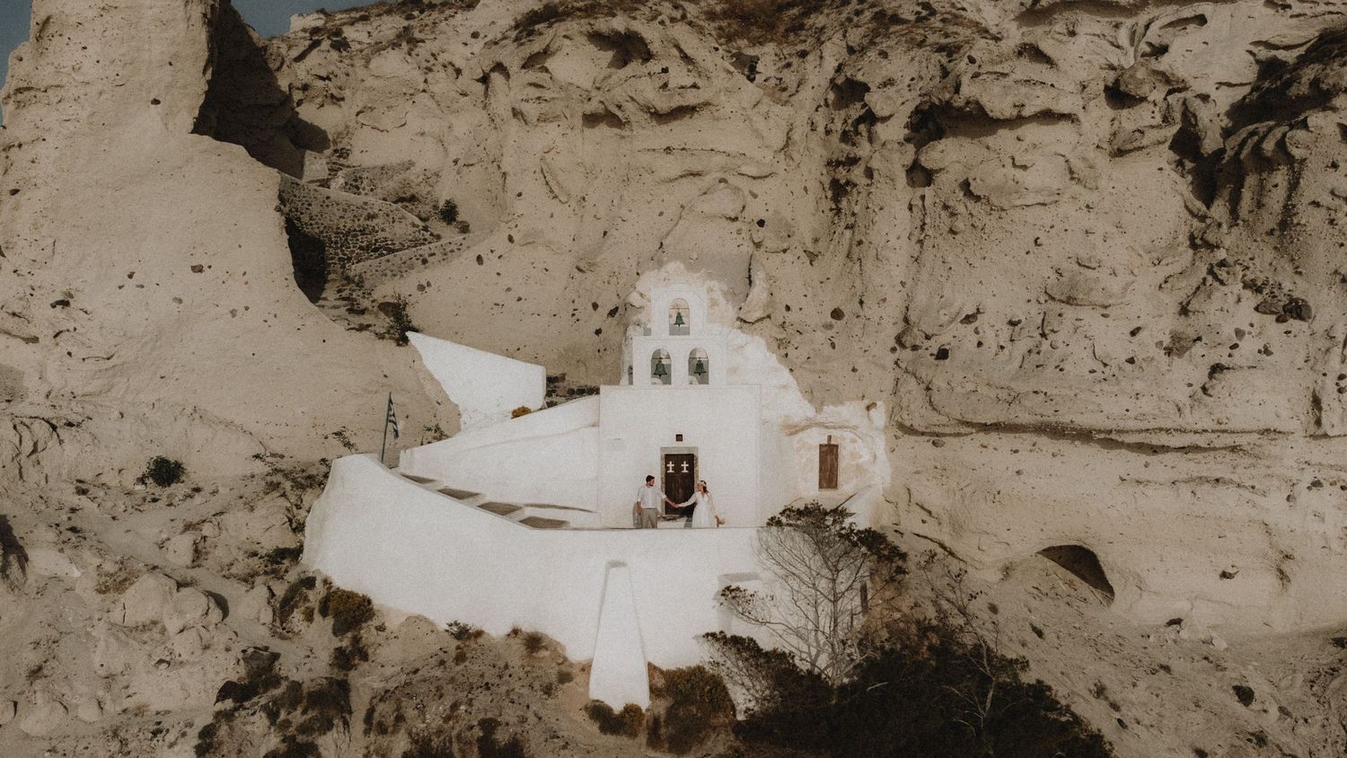 drone wedding photo of a couple in front of a chapel hanging on a cliff during their wedding in Santorini, Greece islands.