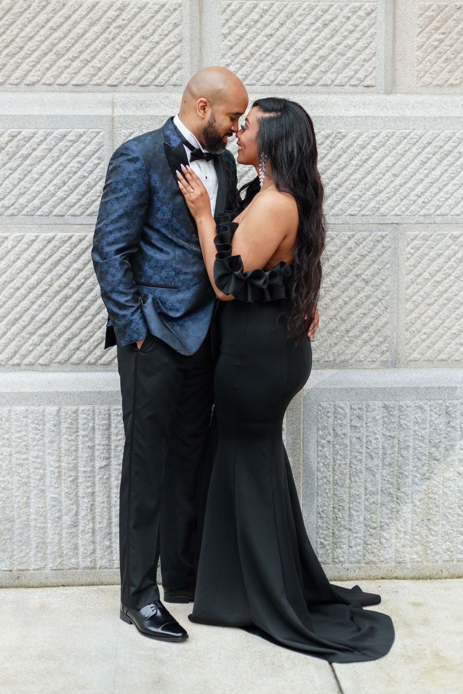 A formal couple in elegant black attire poses against a textured stone wall.