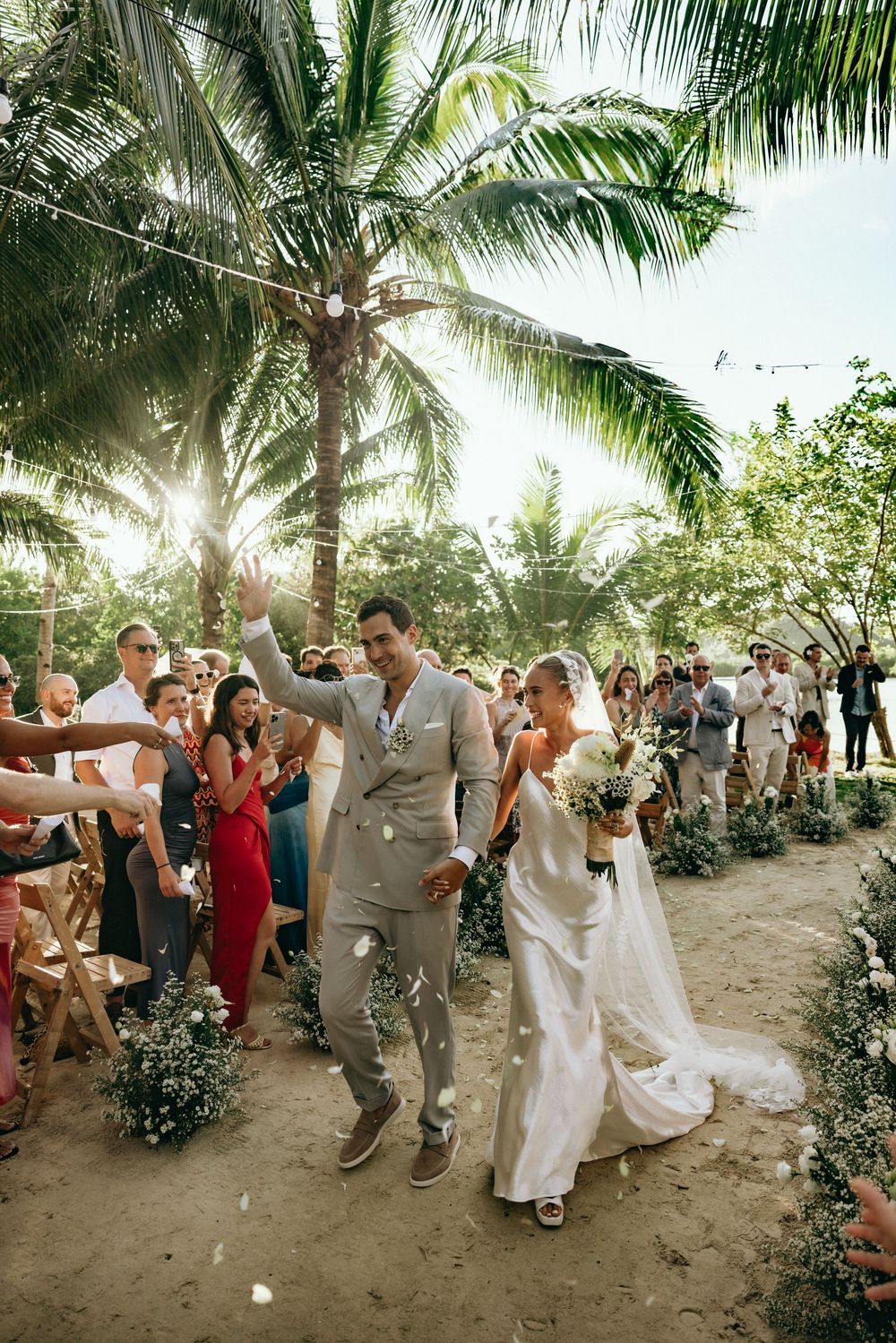Wedding celebration with guests dancing and celebrating on a sandy beach path under palm trees.