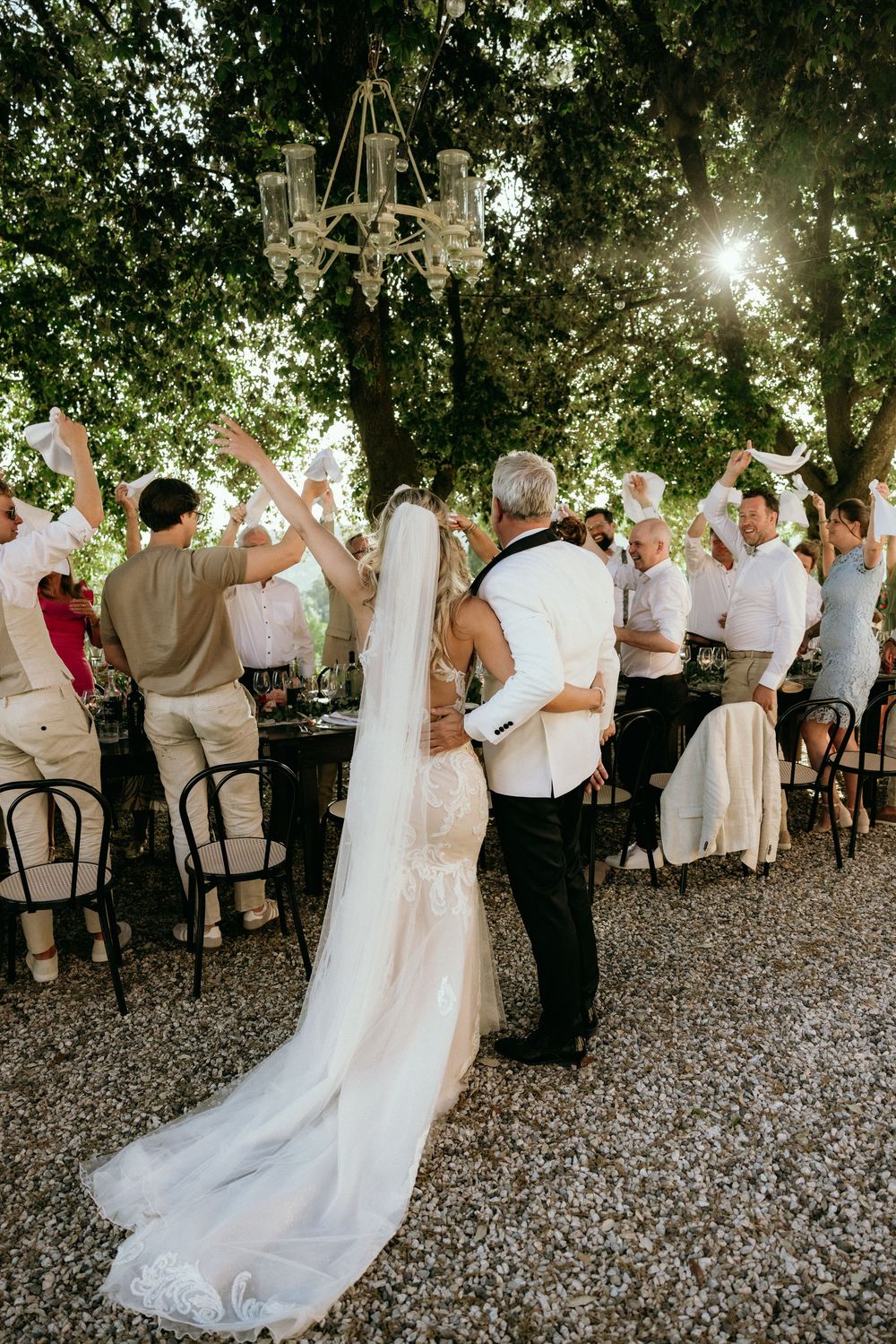 Wedding party dancing in celebration under tree canopy at outdoor reception.