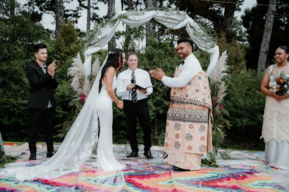 Wedding ceremony under white arch with colorful tie-dye blanket and traditional Pacific Islander officiant.