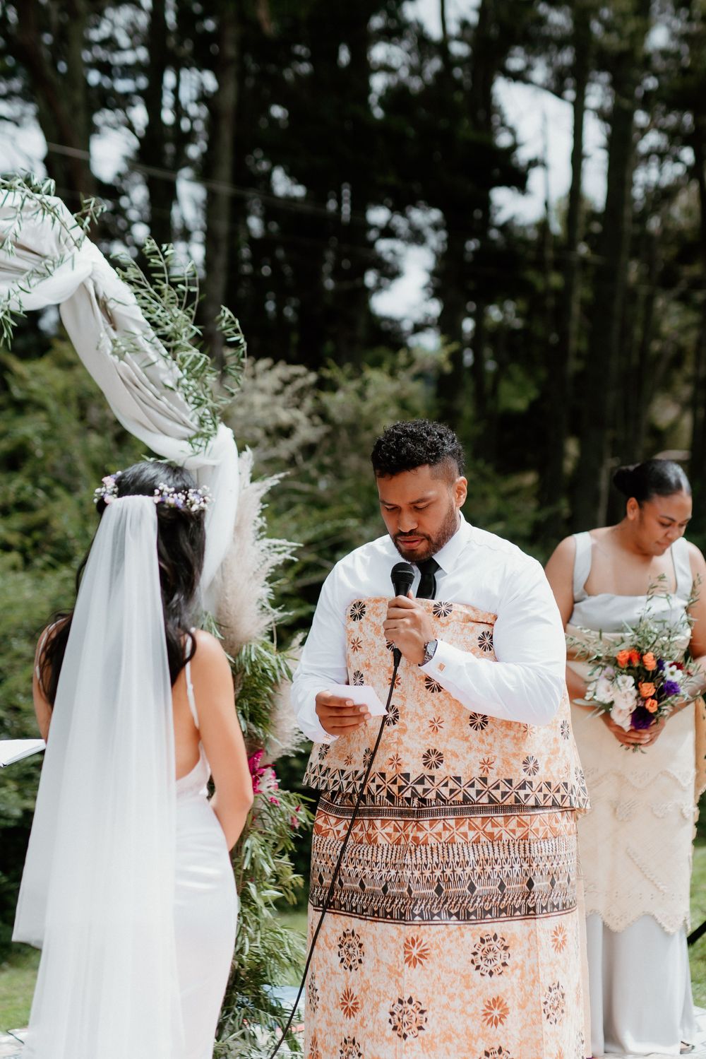 Wedding officiant in traditional patterned robe reads ceremony notes outdoors.