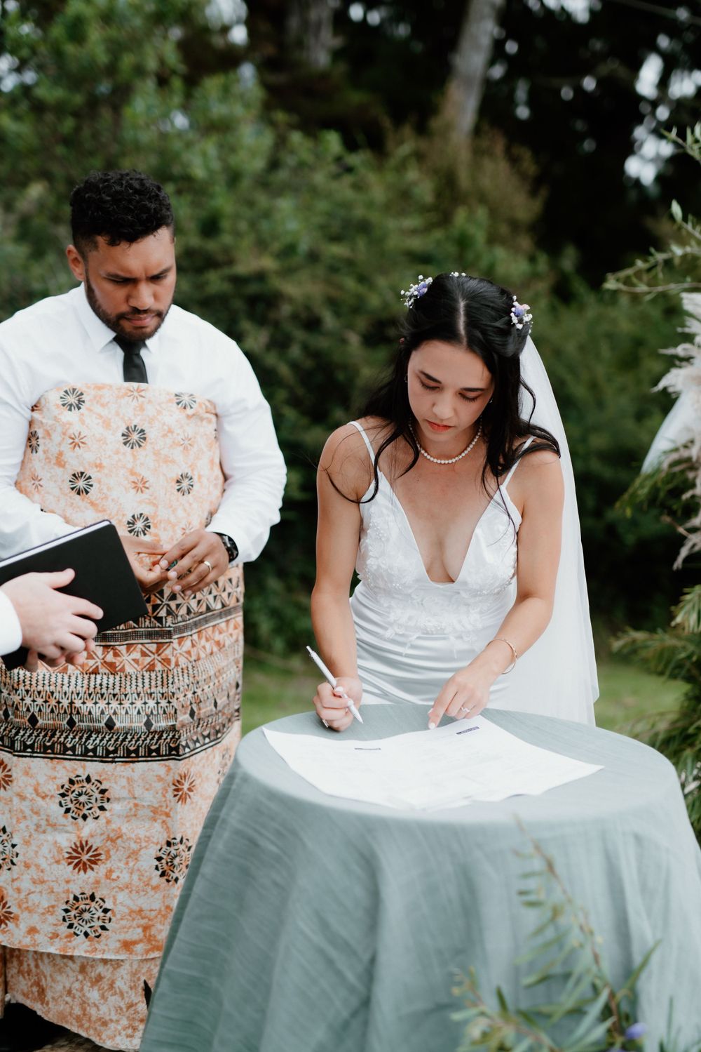 Newlyweds sign marriage documents at an outdoor ceremony with decorative tablecloth.