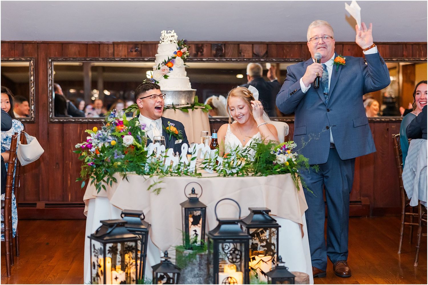 Wedding reception toast with couple at sweetheart table surrounded by decorative lanterns.