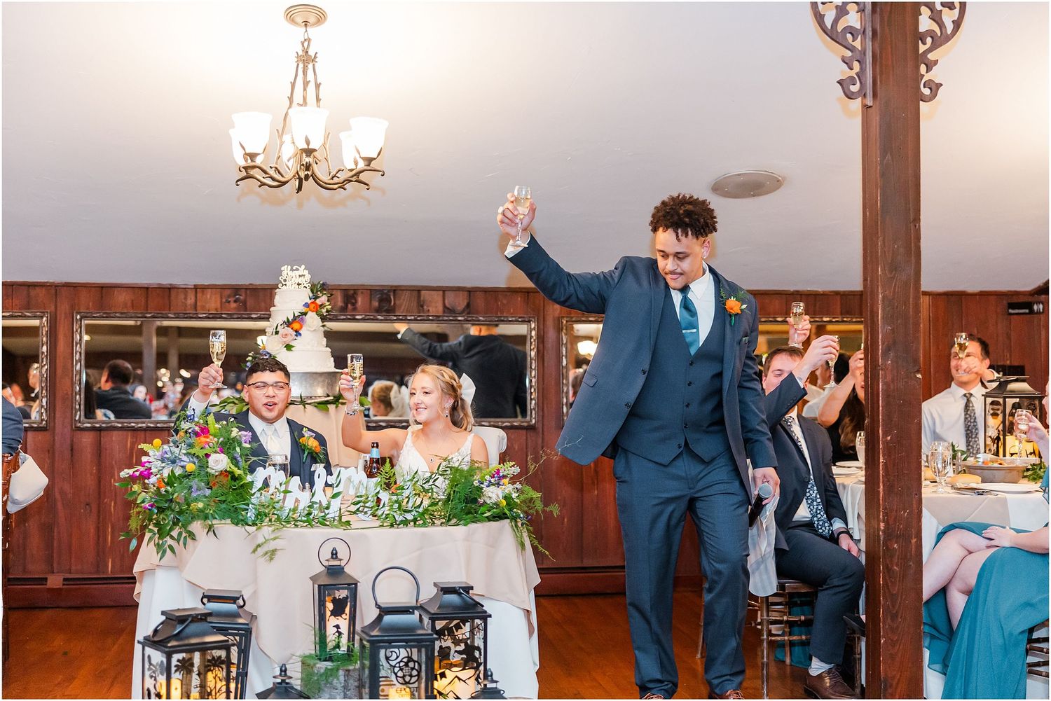 Groom raises glass in celebration at sweetheart table decorated with lanterns and greenery.
