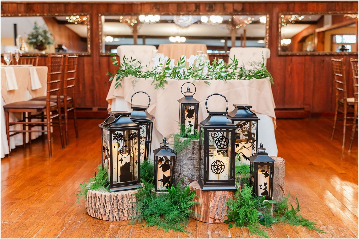 Rustic wedding decor featuring black lanterns arranged on wood slices with greenery accents on a reception table.