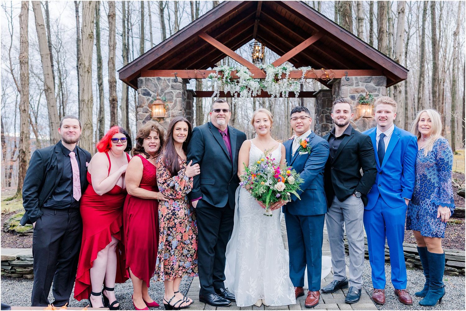 Wedding celebration group photo with guests in colorful attire at a rustic stone venue.