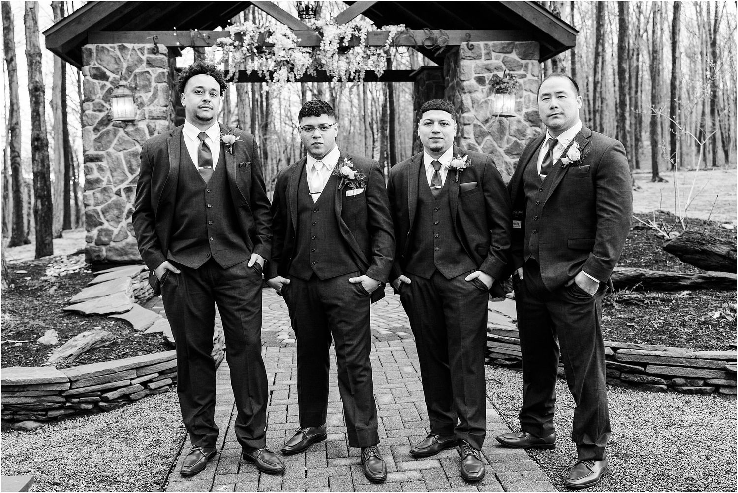 Black and white photo of groomsmen in classic suits standing in front of a stone structure.
