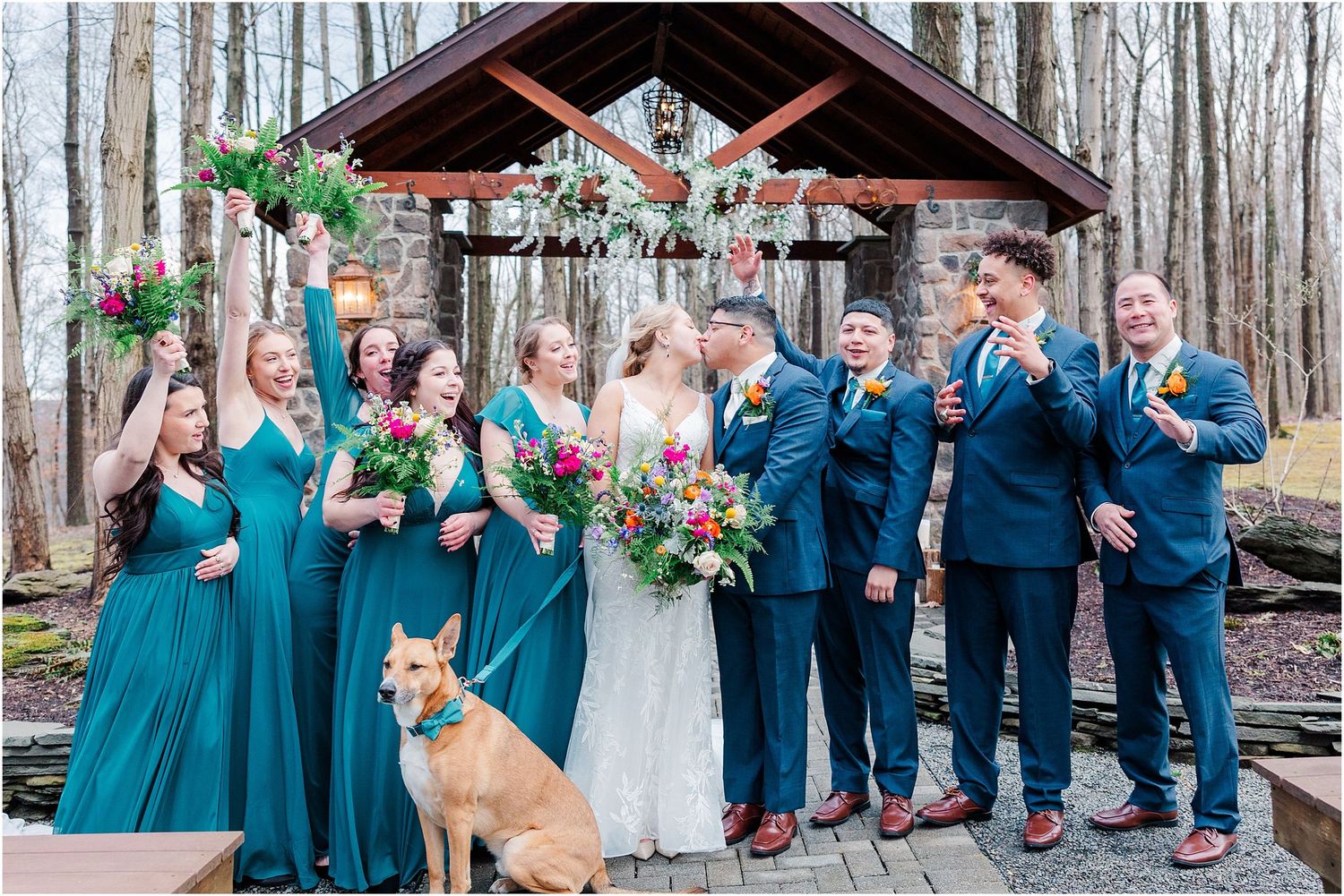 Wedding party in teal dresses and navy suits posing with an adorable dog in front of a rustic stone pavilion.