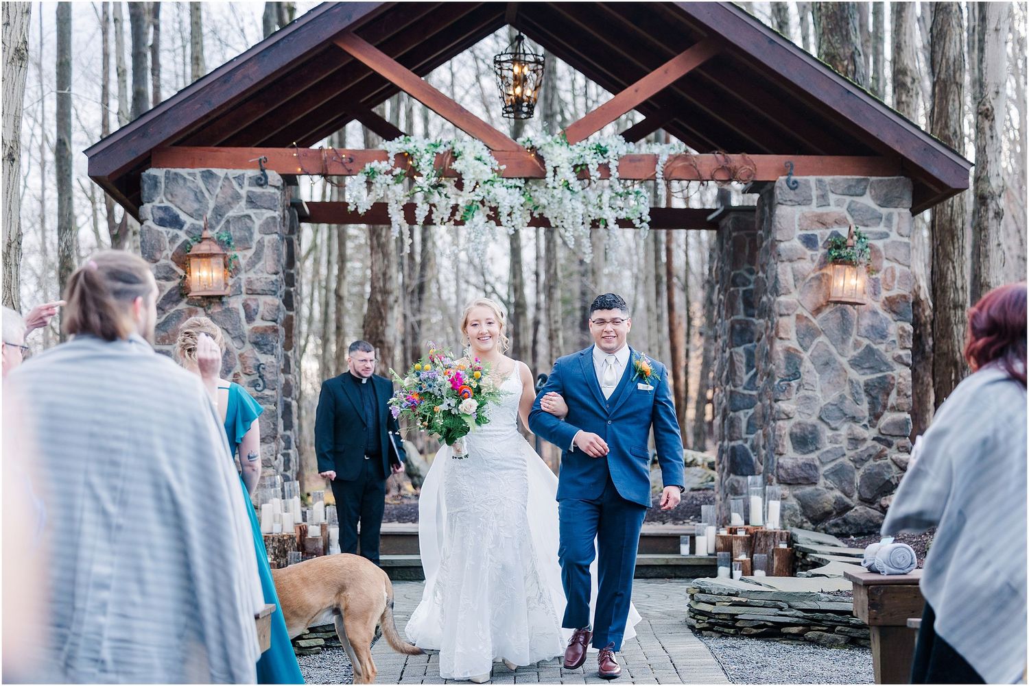 Outdoor wedding ceremony at stone chapel with bride and groom surrounded by guests and dog.