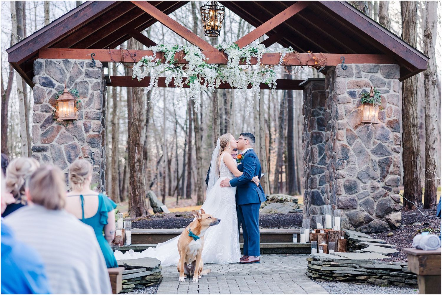 Newlyweds share first kiss at rustic stone chapel while dog watches in foreground.