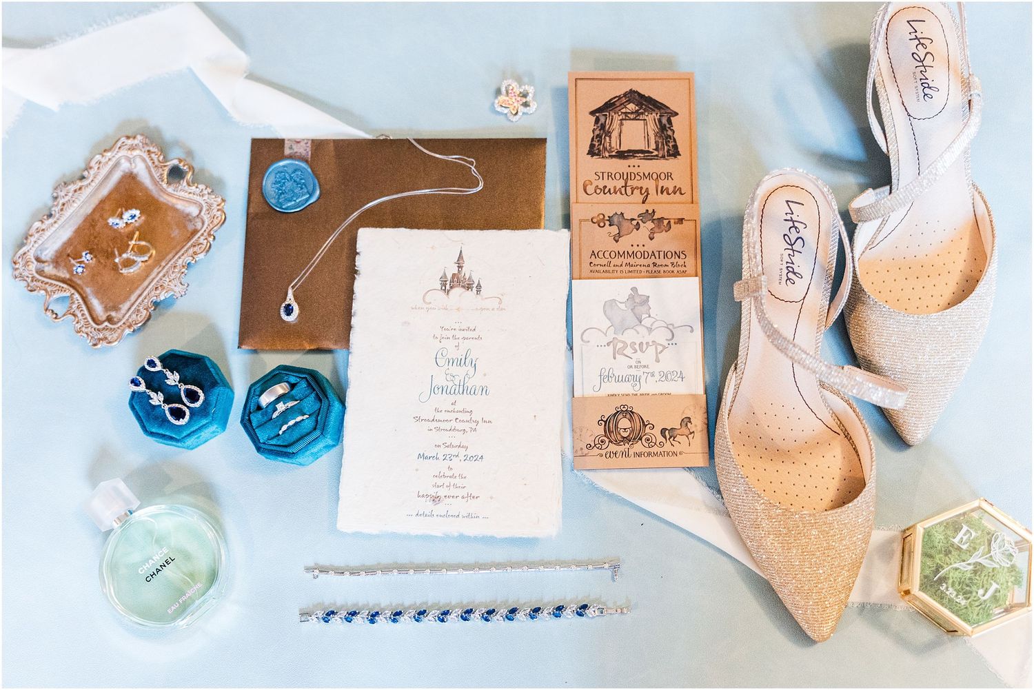 Elegant wedding details including sparkly shoes, invitation, and blue decorative boxes laid out on white surface.