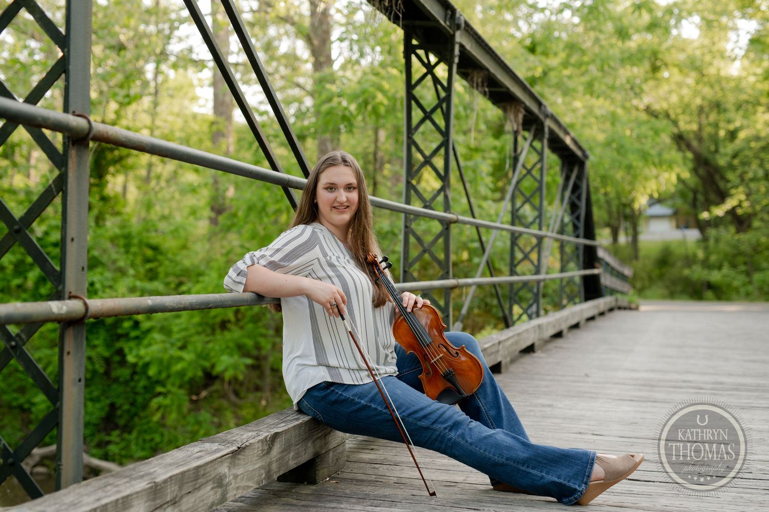 Violinist sitting casually on a rustic wooden bridge surrounded by lush greenery.