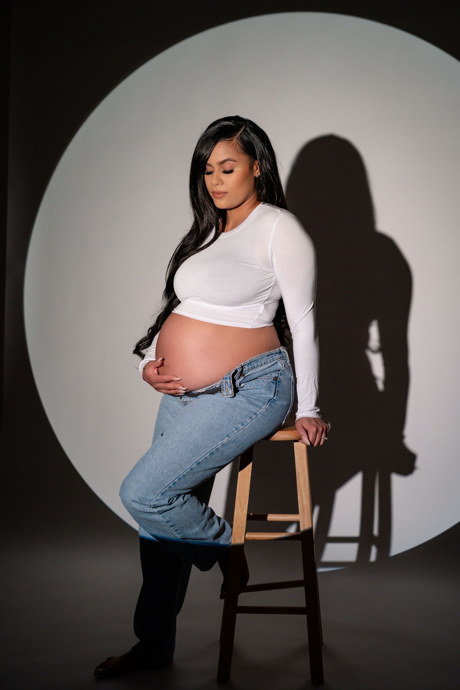 Dramatic studio maternity photos with spotlighting show a pregnant figure in jeans and white top against dark background.