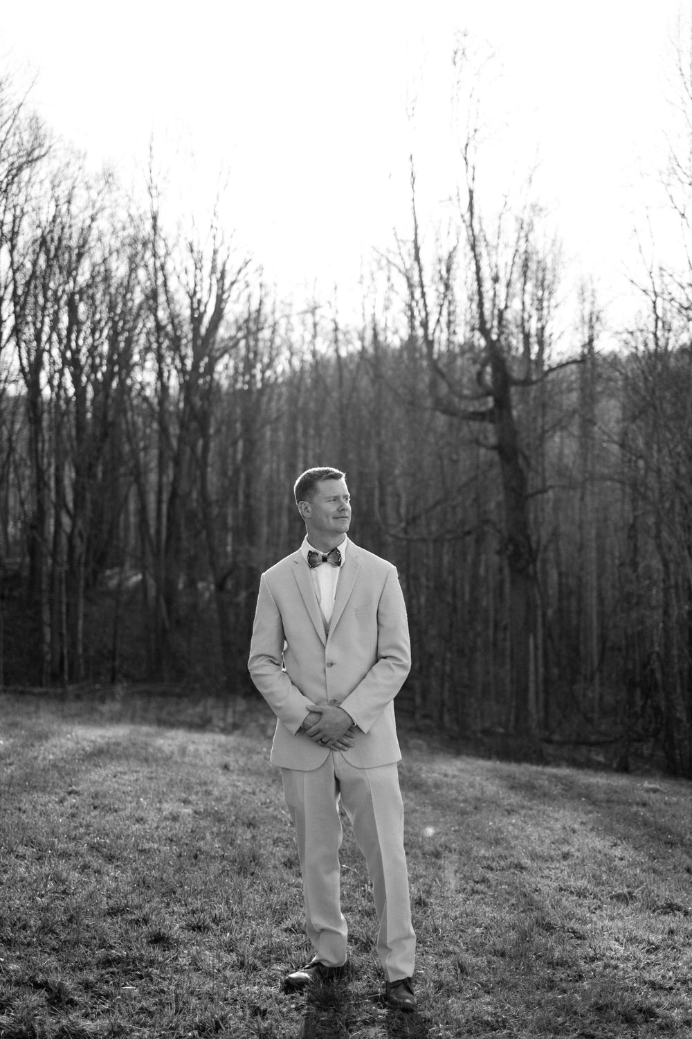Black and white portrait of groom in light suit standing in mountain clearing.