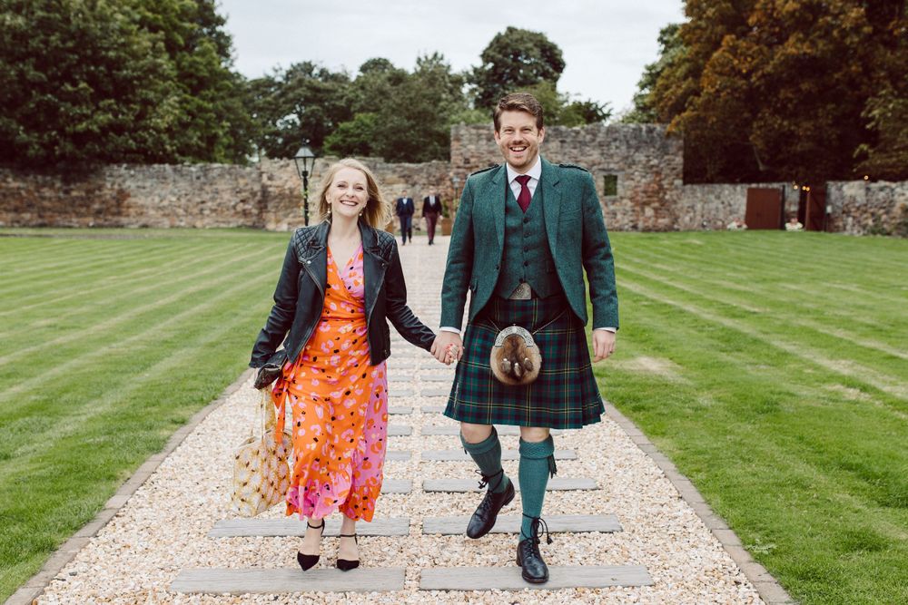 A couple walks along a garden path, with the person on the right wearing a traditional green kilt and sporran.