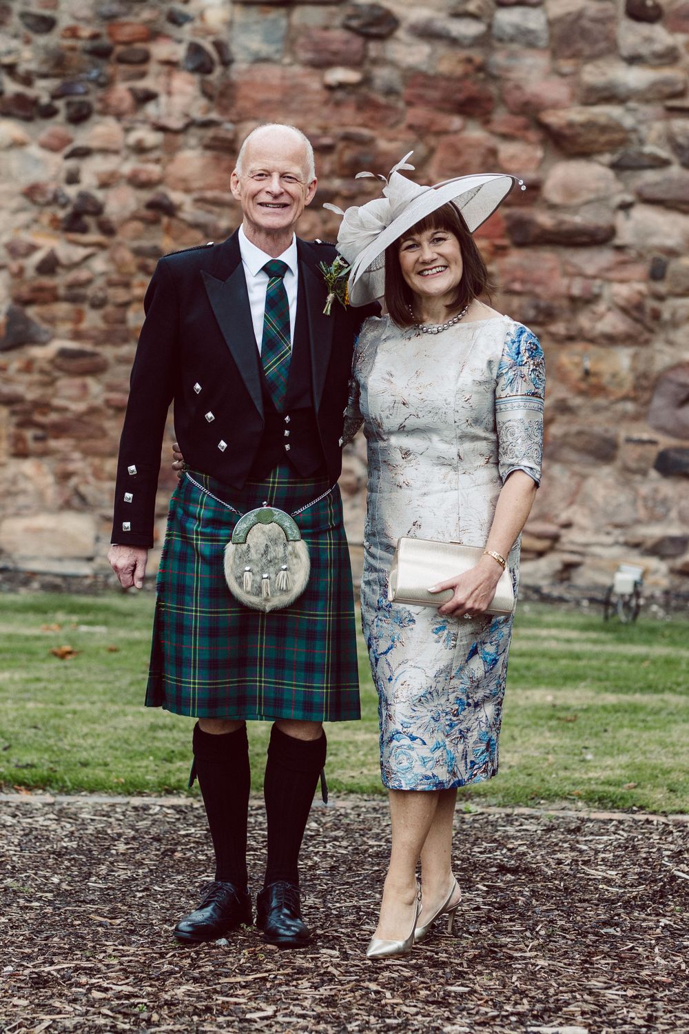 A couple in formal Scottish attire poses against a stone wall at an outdoor event.