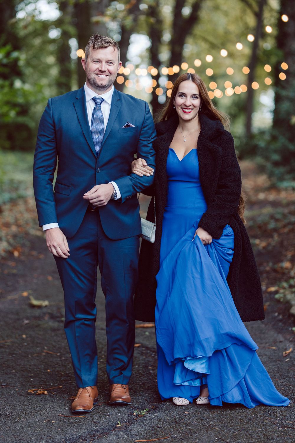A couple poses outdoors at dusk, with the woman wearing a flowing blue gown and black coat.