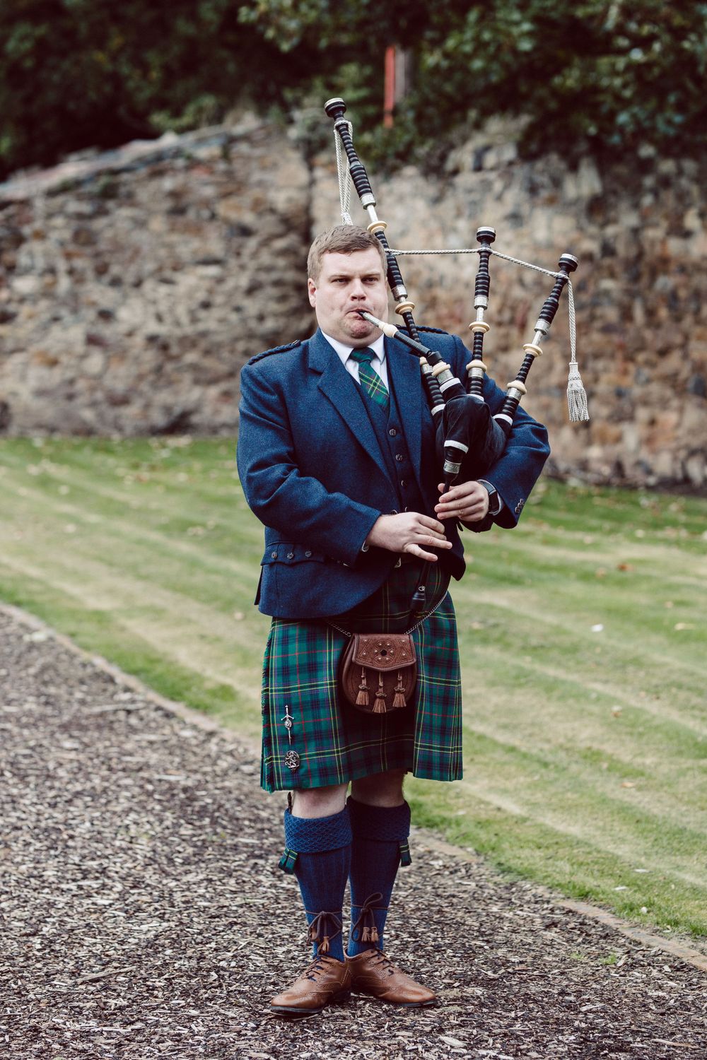 A bagpiper in formal Scottish attire performs outdoors against a stone wall backdrop.