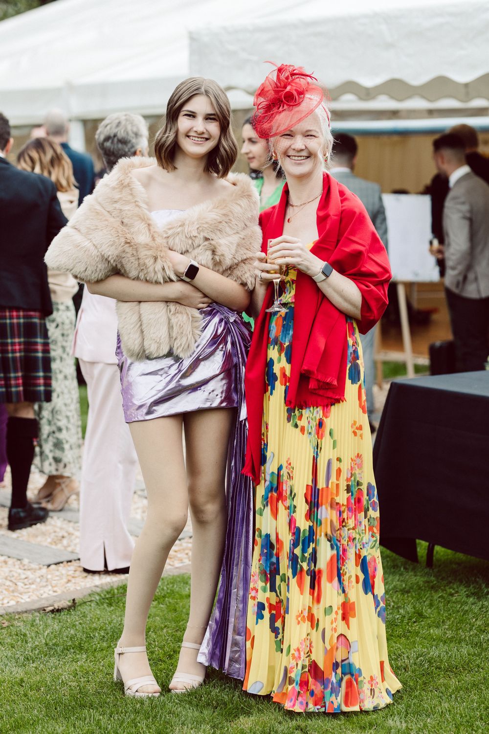 Two guests at an outdoor event wearing colorful formal attire and standing by a white tent.