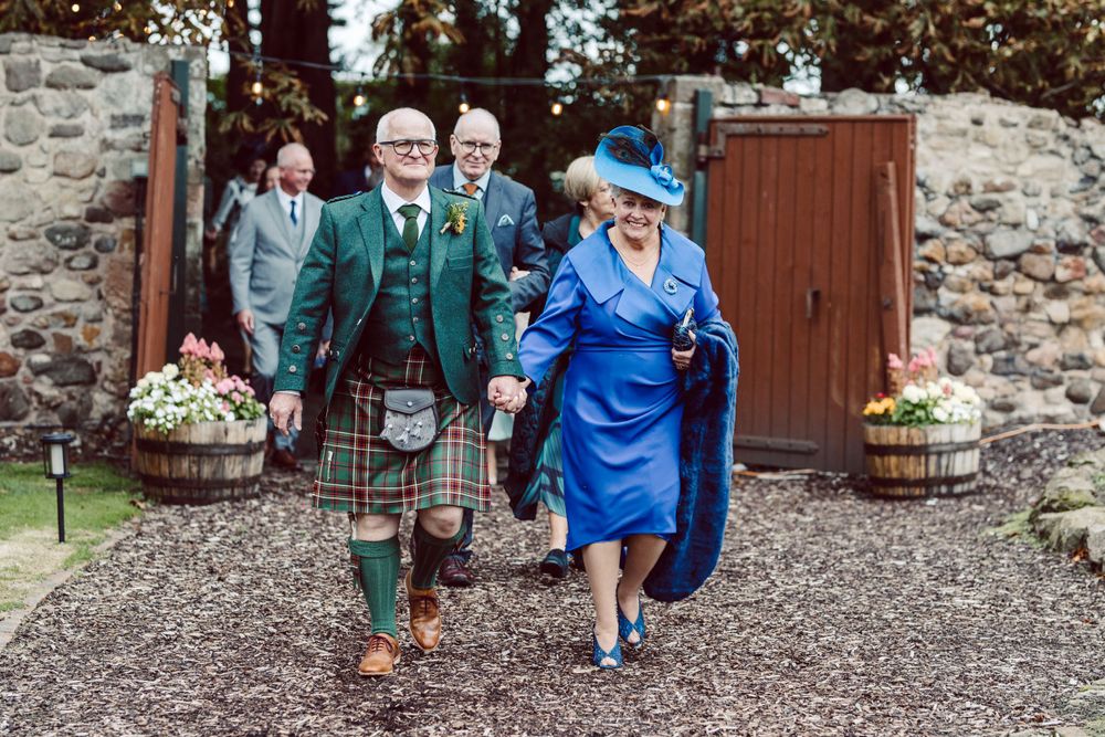 A group walks along a garden path wearing traditional Scottish formal wear and bright blue attire.