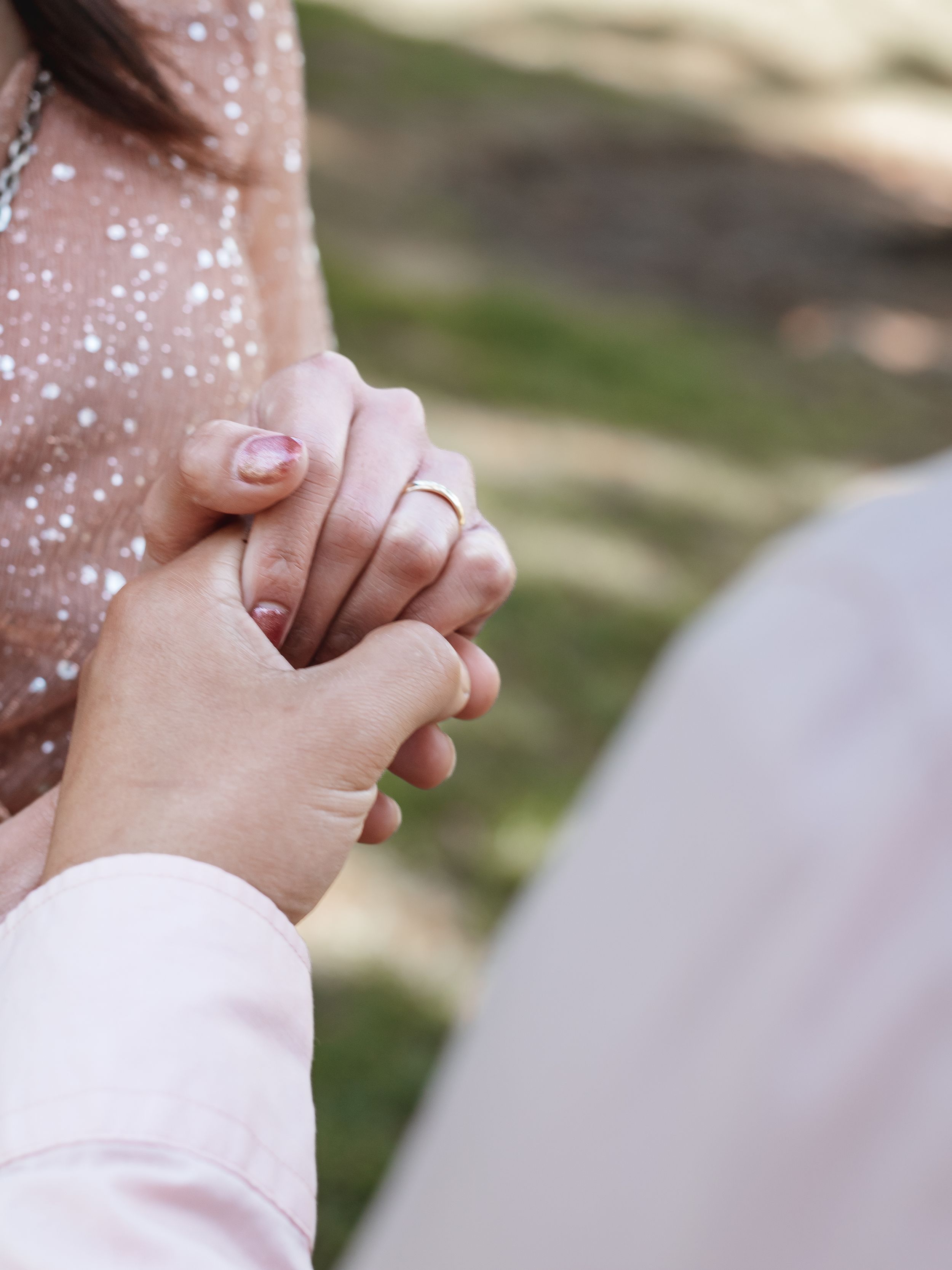 Two hands tenderly clasp together with one wearing a sparkly pink outfit in a romantic outdoor setting.