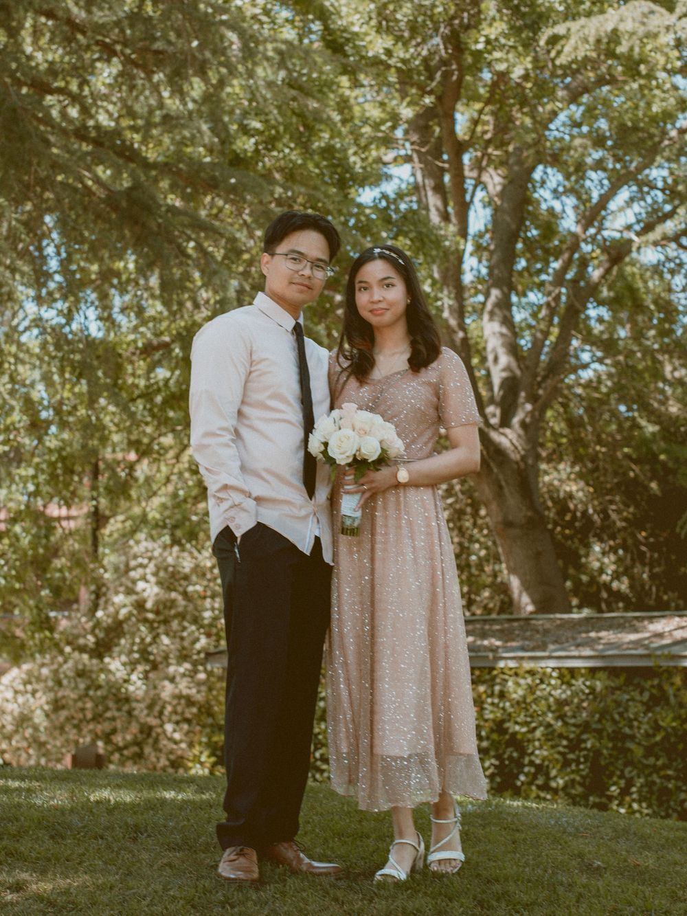 A romantic couple poses outdoors in formal attire with the bride holding a white bouquet.