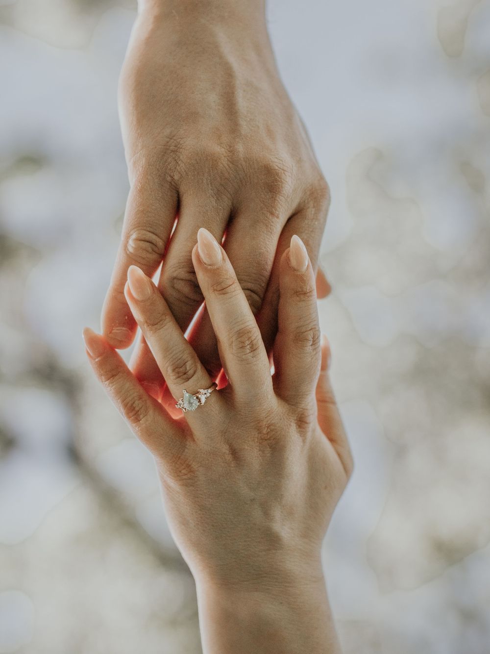 Close-up of two hands wearing engagement ring intertwined against blurred background.