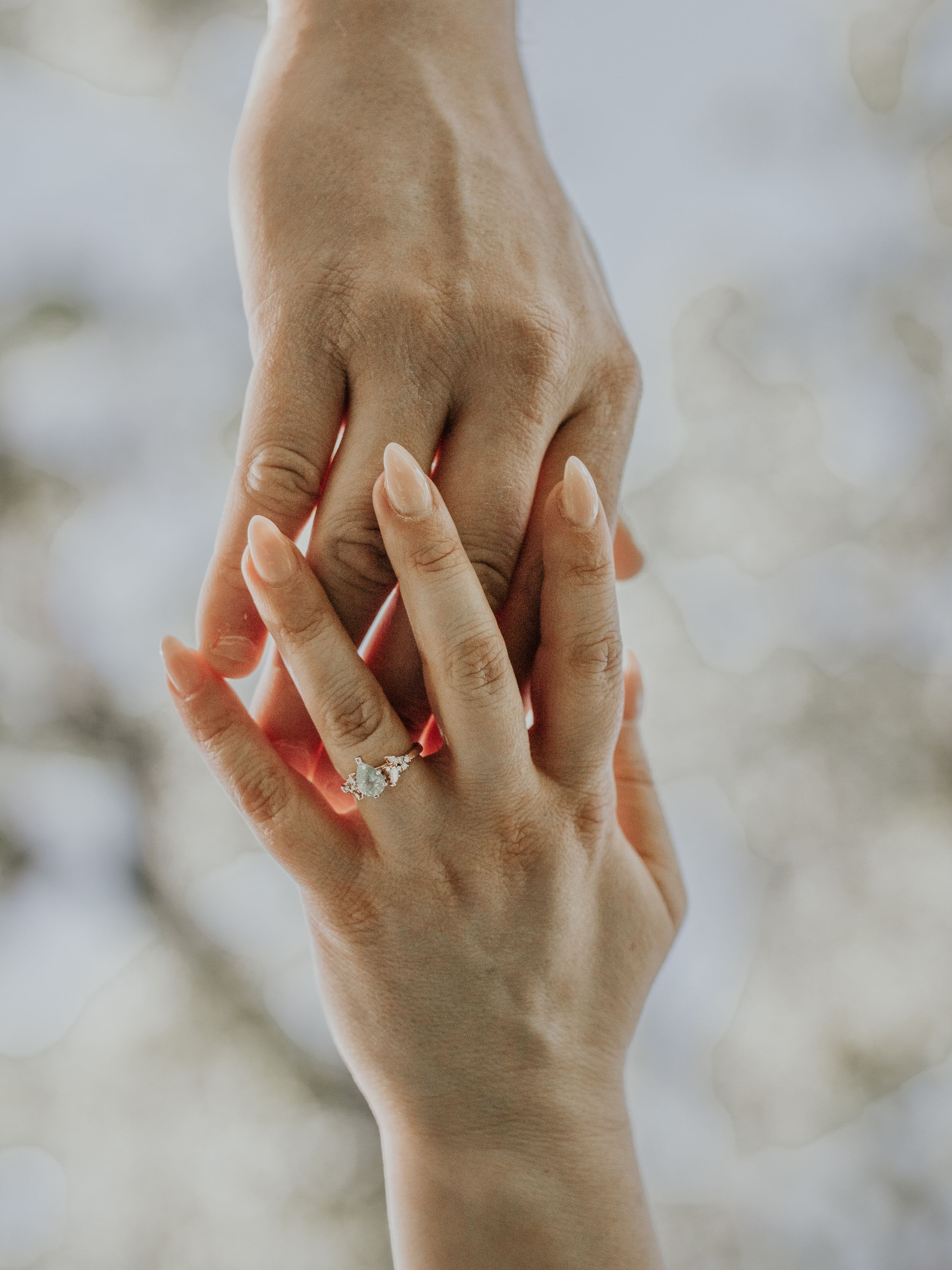 Close-up of two hands wearing engagement ring intertwined against blurred background.