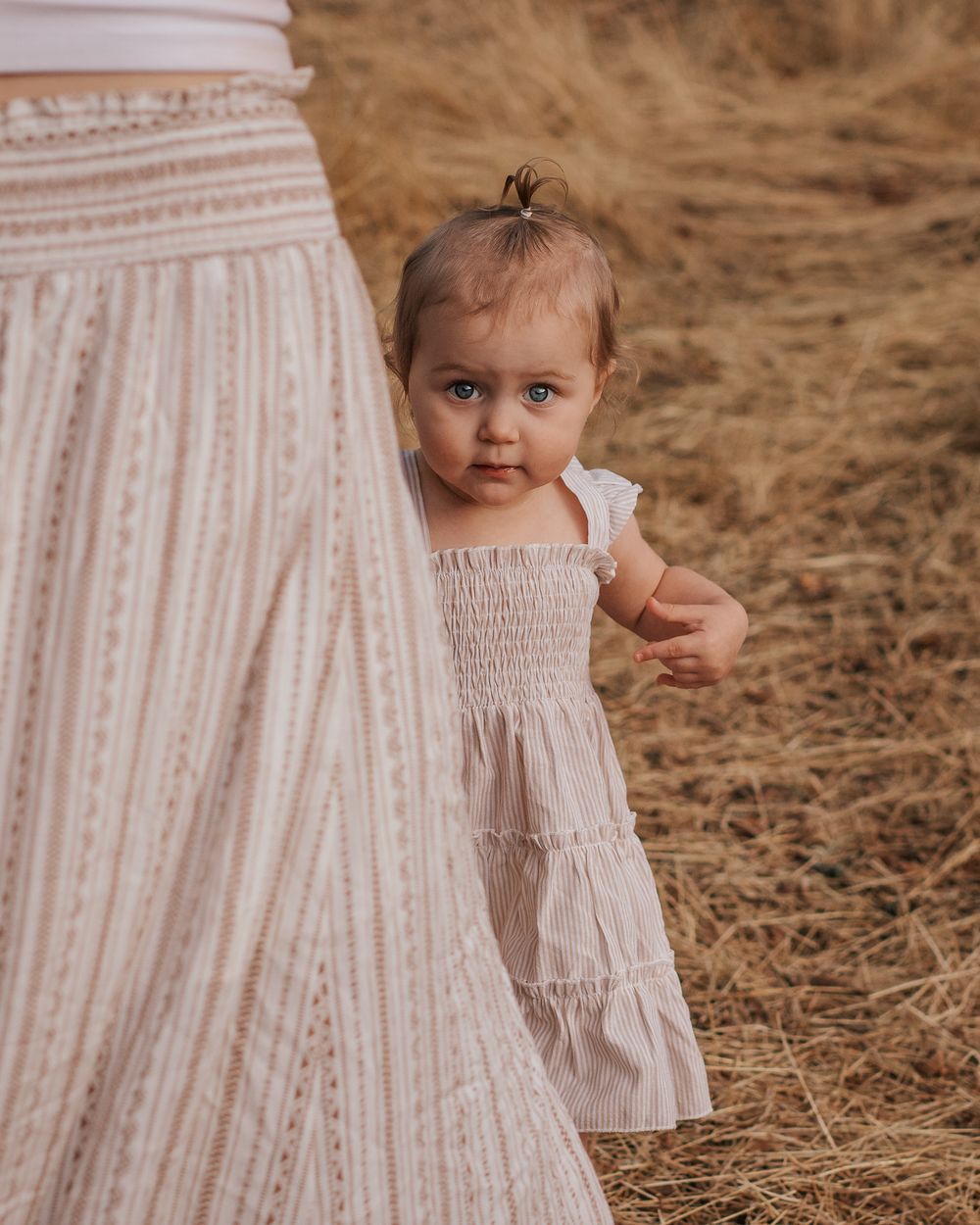 Young child in white dress peeks out from behind parent's skirt in outdoor setting.