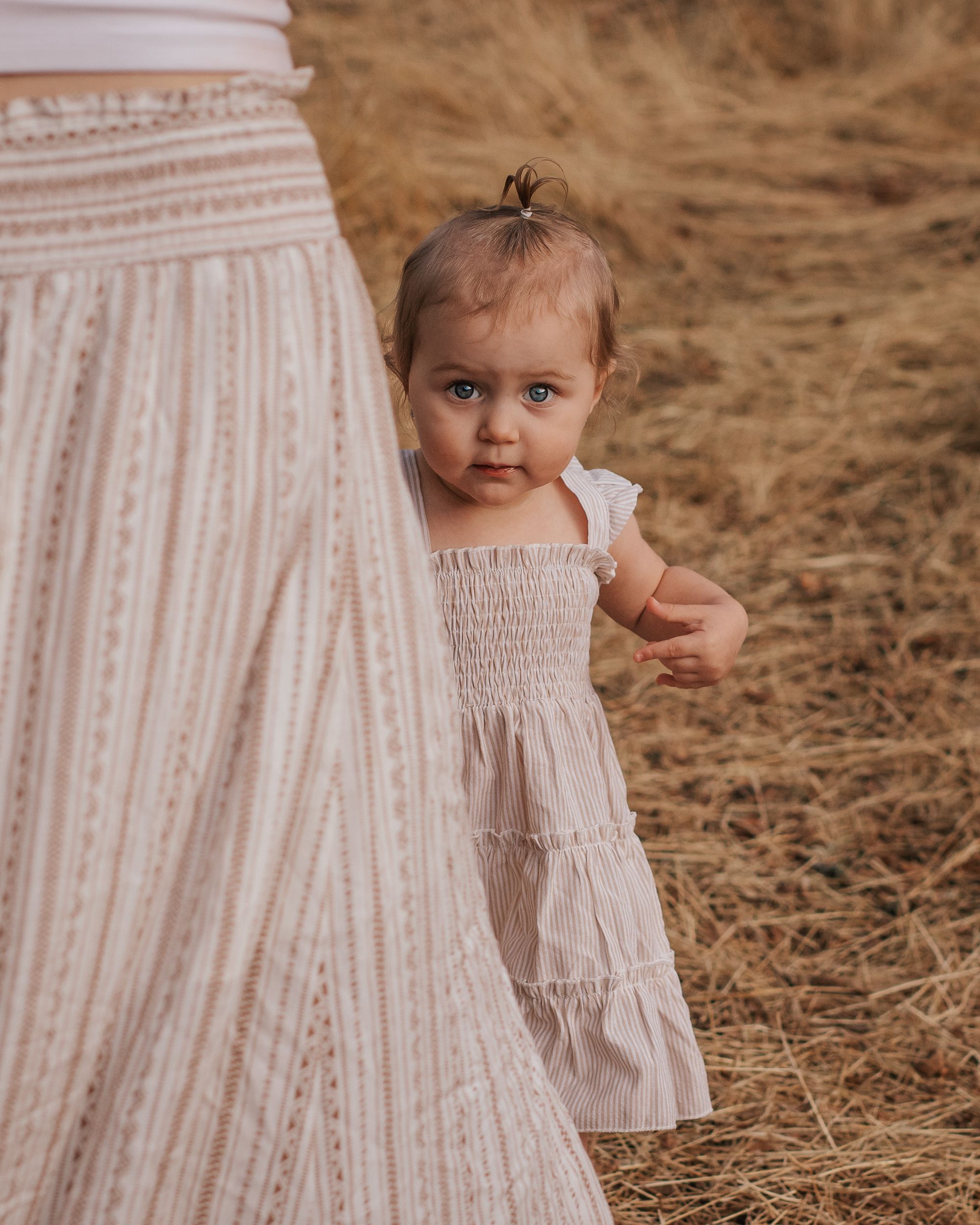 Young child in white dress peeks out from behind parent's skirt in outdoor setting.