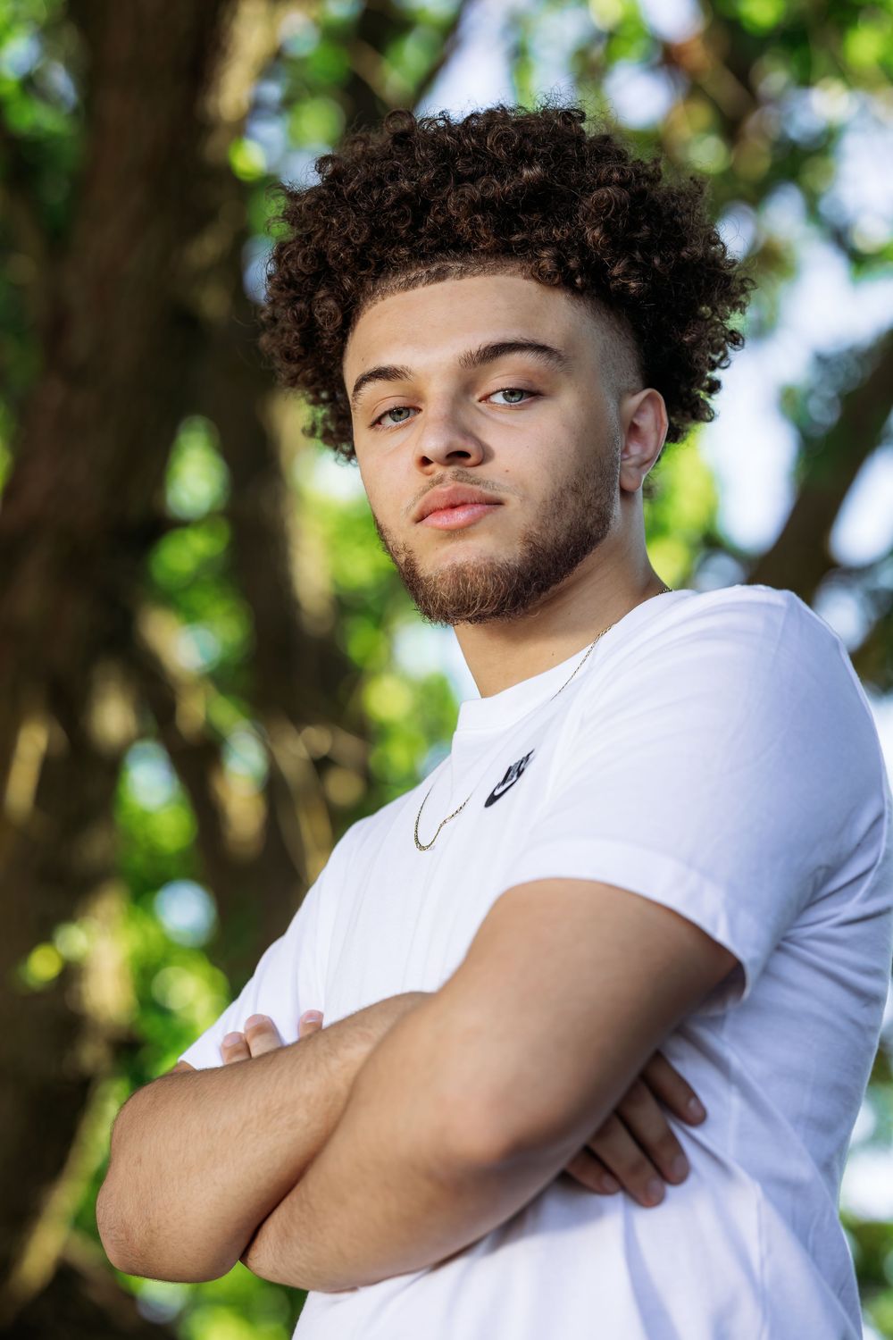 Young person in white t-shirt poses confidently with arms crossed against blurred green outdoor background.
