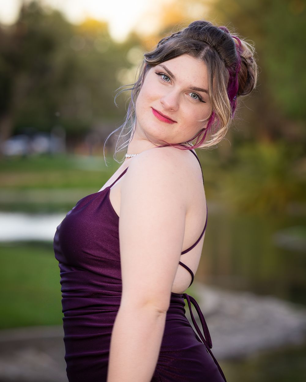 A woman in a dark purple dress poses outdoors at golden hour with soft natural lighting.