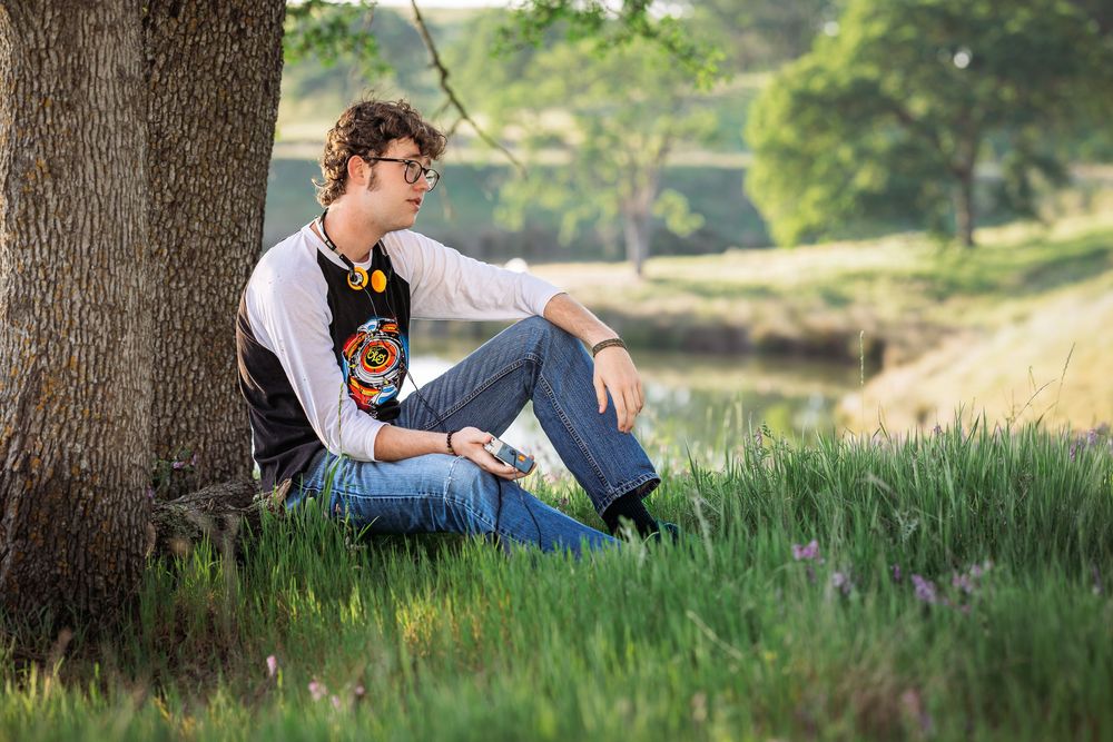 Young person with curly hair sitting against a tree in a lush green park, reading a book on a sunny day.