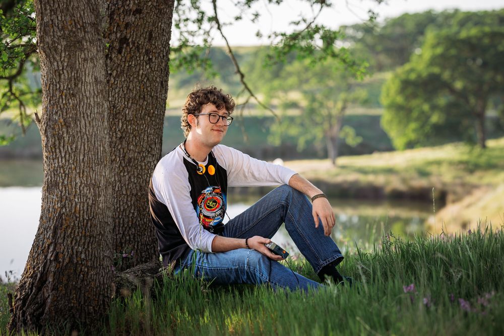 Young man with glasses sitting against a tree in lush green grass near a lake on a sunny day.