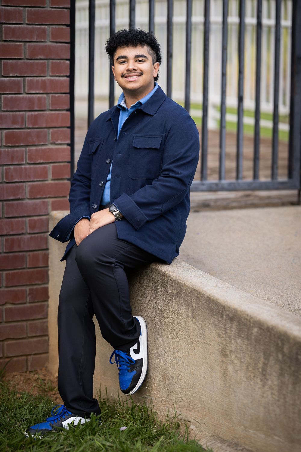 Smiling young man in navy jacket, dark pants, and blue Nike sneakers sitting on a concrete ledge near a brick wall.