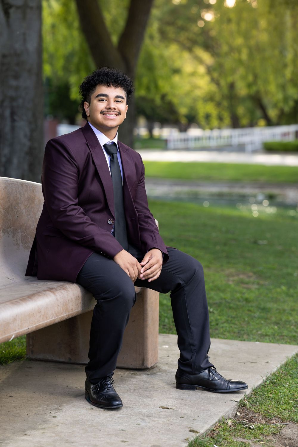 Young man in maroon suit sitting on bench in park, smiling confidently in formal attire.