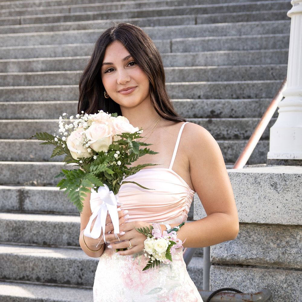 Young woman in pink dress holding white rose bouquet on stone steps, wearing wrist corsage.