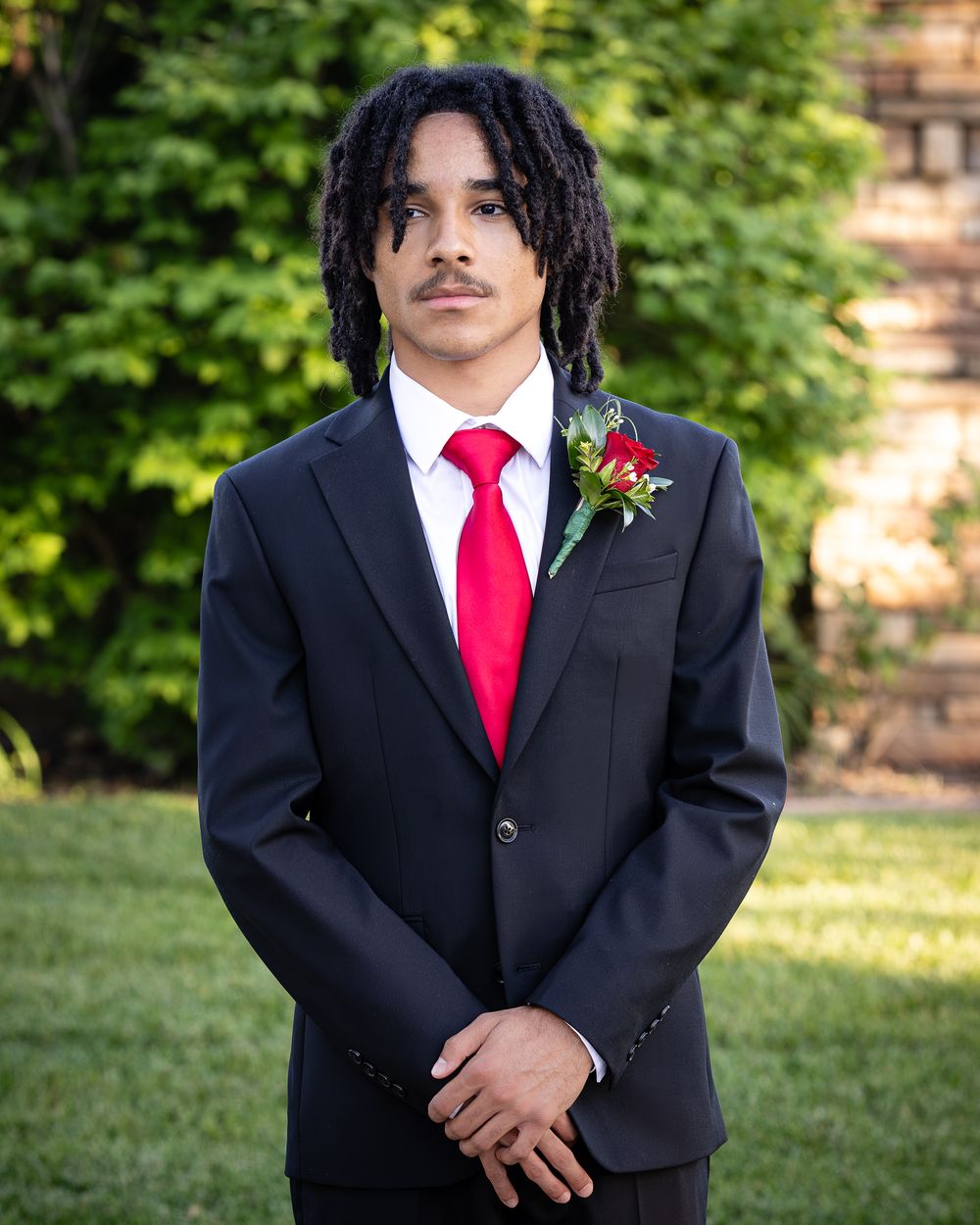 Young man in black suit with red tie and boutonniere poses outdoors for formal prom portrait photo.