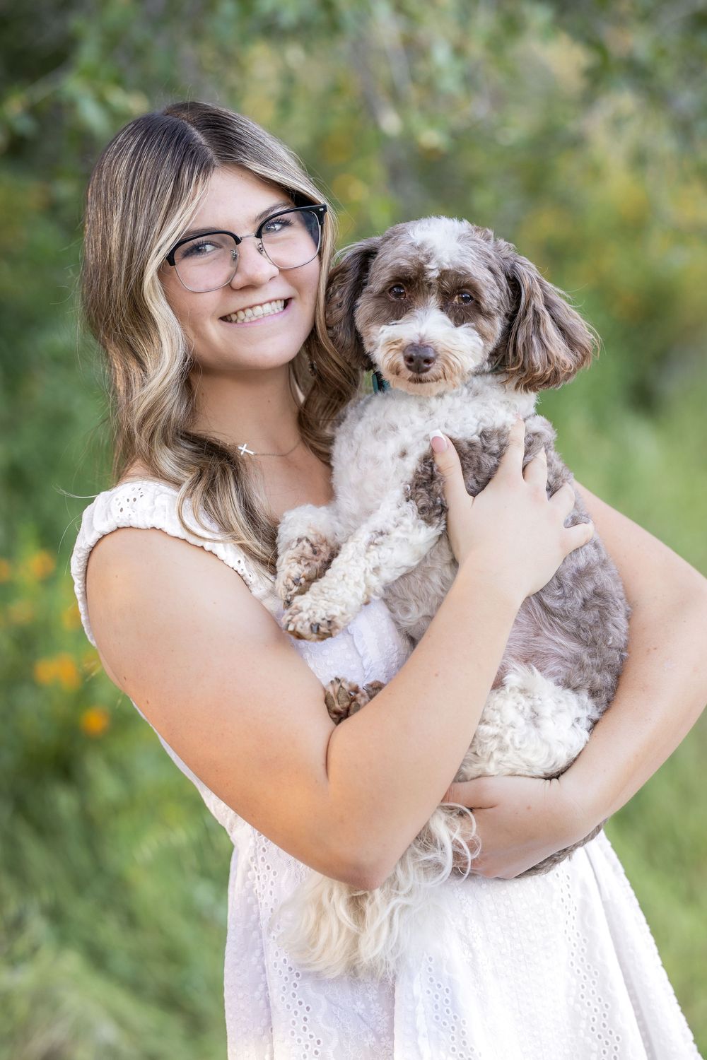 Smiling young woman with glasses holding a fluffy brown and white dog outdoors in a green garden setting.