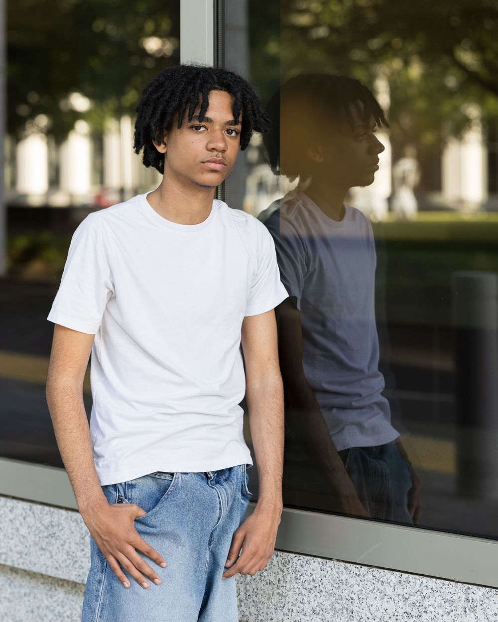 Young man in white t-shirt and jeans leaning against glass wall with reflection visible outdoors.
